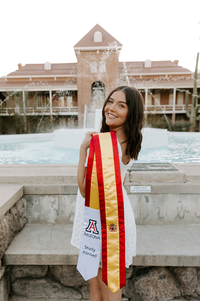 Madison in white dress in front of Old Main Fountain, with yellow and red Study Abroad graduation stole draped over left shoulder