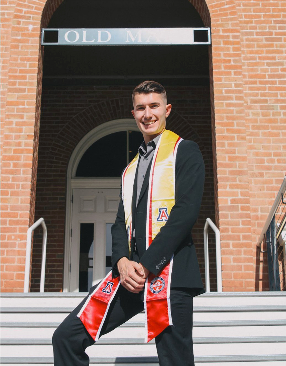 Luke Hoeferkamp in a dark grey suit with graduation stoles in red and yellow in front of Old Main