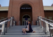 International student Veronica sits on steps at Old Main, wearing graduation stole and holding cap