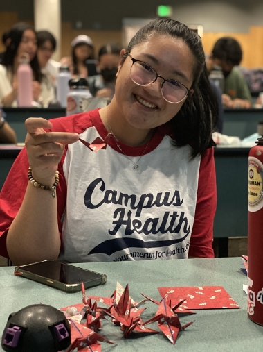 Student wearing a red and white t-shirt with the words "Campus Health." They are holding a paper crane they have made, with several more on the table.
