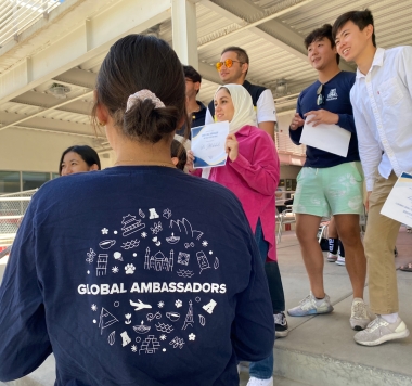 GA Shows back of blue Global Ambassadors tshirt with icons representing cultures and travel, other students in background