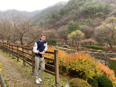 Luke Hoeferkamp leans on a wooden fence in the countryside in S. Korea. It is fall and the leaves are changing colors.